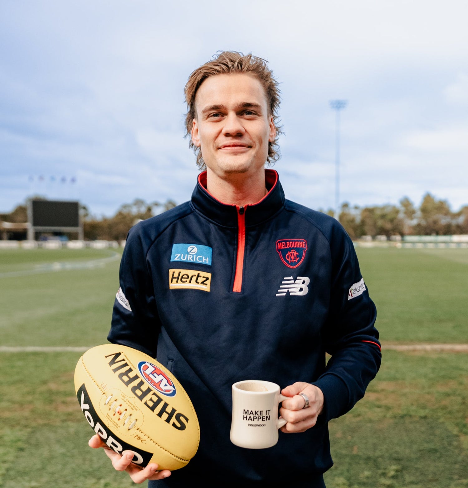 Melbourne Football Club player, Trent Rivers, holding a football and a mug on a sports field